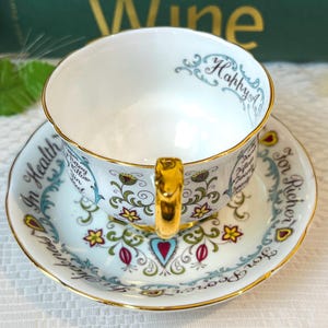 May include: A white teacup and saucer set with gold trim and floral designs. The teacup has the words "Happy" and "In Health" written on it. The saucer has the words "For Richer" and "For Poorer" written on it.