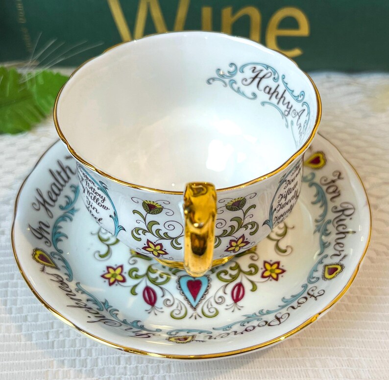 May include: A white teacup and saucer set with gold trim and floral designs. The teacup has the words "Happy" and "In Health" written on it. The saucer has the words "For Richer" and "For Poorer" written on it.
