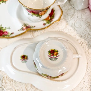 May include: A white teacup and saucer with a floral pattern. The teacup has a gold rim and the saucer has a scalloped edge. The bottom of the teacup has the words "Old Country Roses" and "Royal Albert Bone China England".