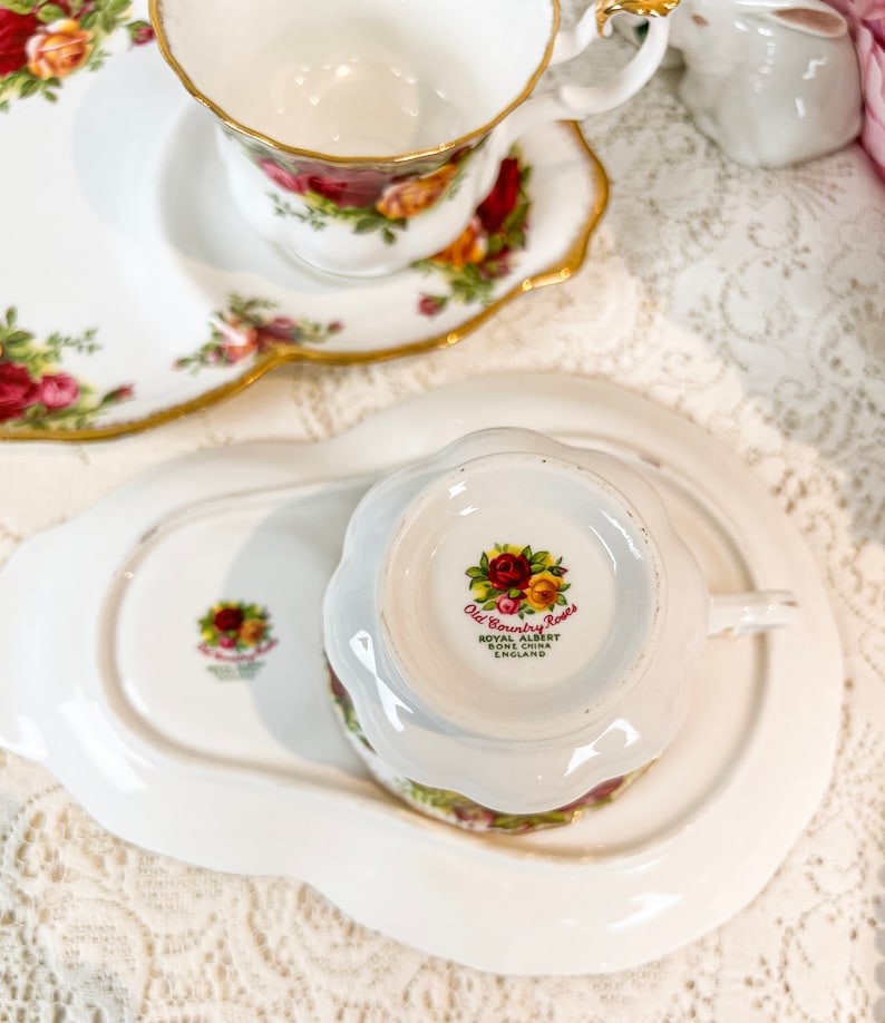 May include: A white teacup and saucer with a floral pattern. The teacup has a gold rim and the saucer has a scalloped edge. The bottom of the teacup has the words "Old Country Roses" and "Royal Albert Bone China England".