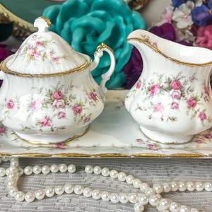 May include: A white porcelain sugar bowl with a lid and a matching creamer pitcher, both featuring pink rose floral patterns and gold trim. These two pieces sit on a rectangular porcelain tray with matching floral designs and gold edges.