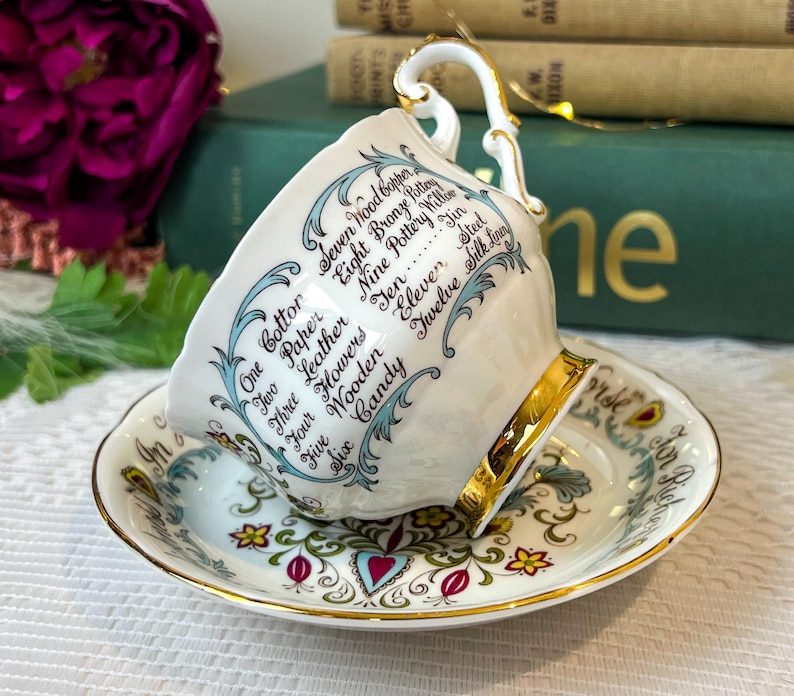 May include: A white teacup and saucer with a gold trim. The teacup has a blue and white floral design and the words "One Cotton, Two Paper, Three Leather, Four Flowers, Five Wooden, Six Candy, Seven Wood Copper, Eight Bronze Pottery, Nine Pottery Willow, Ten Jin, Eleven Steel, Twelve Silk Linen" written around the side. The saucer has a similar floral design with a gold trim.