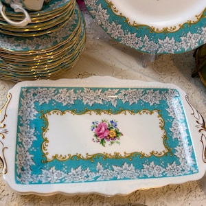 May include: A white rectangular serving tray with a turquoise and gold border. The border is decorated with white floral designs. The centre of the tray features a white rectangle with a gold border and a floral design in pink, yellow, and purple.