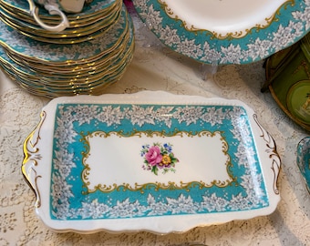 May include: A white rectangular serving tray with a turquoise and gold border. The border is decorated with white floral designs. The centre of the tray features a white rectangle with a gold border and a floral design in pink, yellow, and purple.