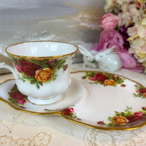 May include: A white teacup and saucer with a gold rim and a floral pattern of red, yellow, and green roses. The saucer is shaped like a plate with a small indentation for the teacup.
