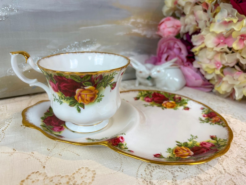 May include: A white teacup and saucer with a gold rim and a floral pattern of red, yellow, and green roses. The saucer is shaped like a plate with a small indentation for the teacup.