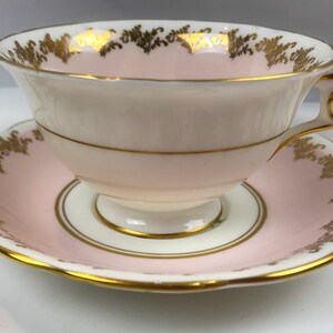 Puede incluir: Un juego de taza y platillo de t&eacute; rosa y blanco con ribetes dorados. La taza tiene un delicado dise&ntilde;o floral y el platillo tiene un borde festoneado.