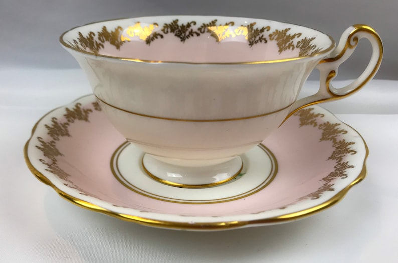 Puede incluir: Un juego de taza y platillo de t&eacute; rosa y blanco con ribetes dorados. La taza tiene un delicado dise&ntilde;o floral y el platillo tiene un borde festoneado.