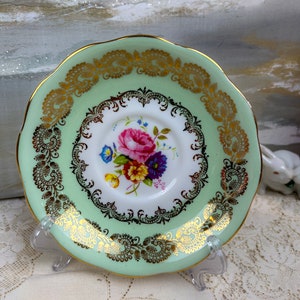May include: A vintage teacup saucer with a light green border and gold filigree detailing. The center features a floral bouquet of pink, purple, yellow, and blue flowers. The saucer is displayed on a clear stand.
