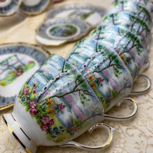 May include: A set of vintage teacups and saucers. The cups and saucers feature a landscape design with birch trees, flowers, and a blue sky. The rims and handles are trimmed with gold. The teacups are stacked in a row.