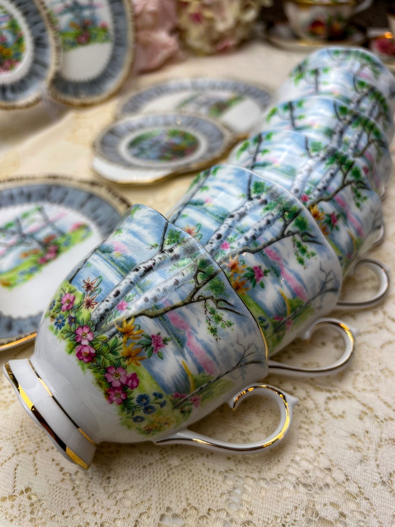 May include: A set of vintage teacups and saucers. The cups and saucers feature a landscape design with birch trees, flowers, and a blue sky. The rims and handles are trimmed with gold. The teacups are stacked in a row.