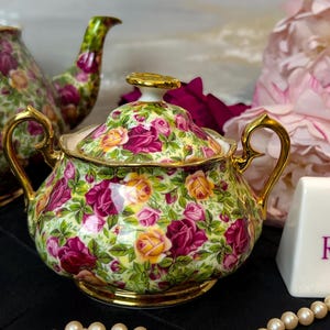 May include: A floral-patterned sugar bowl with a lid, featuring a design of pink and yellow roses on a green background. The bowl has gold handles and trim. A teapot is visible in the background. A white sign with purple text is also present.