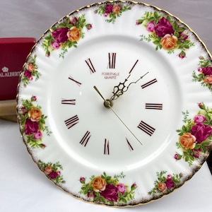 Puede incluir: Un reloj de porcelana blanca con borde dorado y diseño floral. La esfera del reloj tiene números romanos y dice "Forestville Quartz".