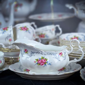 Op de afbeelding: Een set witte porseleinen theekopjes en schoteltjes met een bloemenpatroon. De kopjes en schoteltjes hebben een delicaat, vrouwelijk ontwerp met roze en blauwe bloemen. De set bevat een juskom met een bijpassend bloemenpatroon.