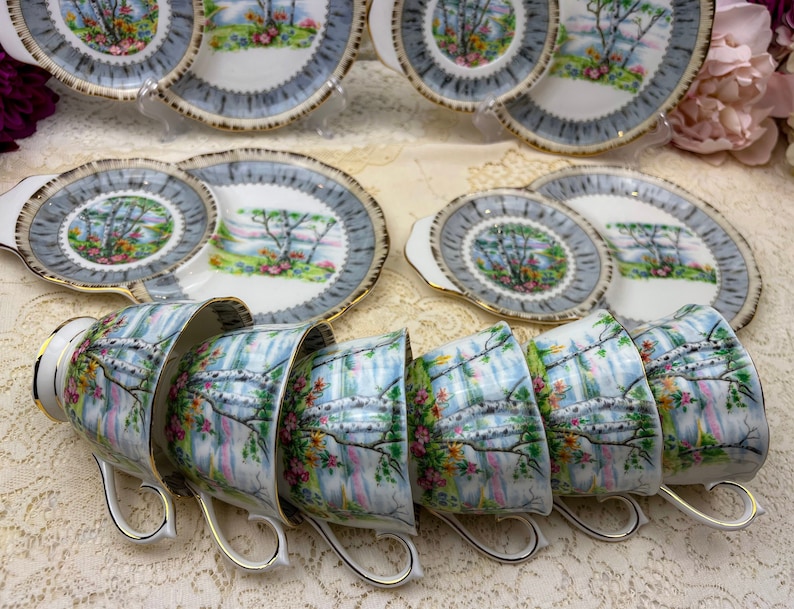 May include: A vintage tea set featuring cups and saucers with a scenic landscape design. The cups and plates have a light blue border and gold trim. The landscape design includes trees, flowers, and a waterfall.