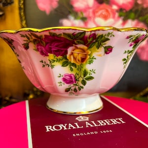 May include: A pink and white porcelain bowl with a gold rim and floral design. The bowl is decorated with a pattern of roses and leaves. The bowl is on a pink and red box with the words "Royal Albert England 1904" printed on it.