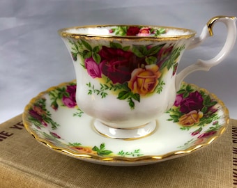May include: A white teacup and saucer with a gold rim and floral pattern. The floral pattern features red, pink, and yellow roses with green leaves.