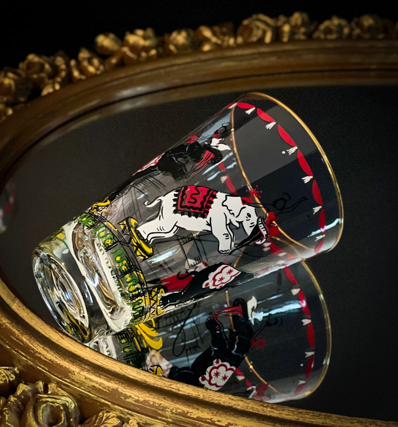 May include: A clear glass tumbler with a circus theme. The tumbler features a black and white illustration of elephants, clowns, and other circus performers. The tumbler has a red and white striped band around the top.