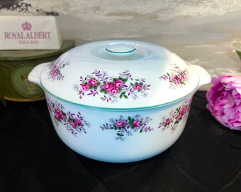 May include: White ceramic casserole dish with a lid, featuring a floral pattern of pink roses and green leaves. The dish has a green trim around the rim.