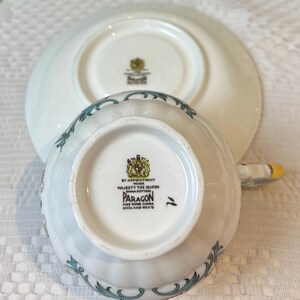 May include: White teacup and saucer with a blue and gold floral pattern. The bottom of the teacup has the text "By Appointment to Her Majesty The Queen China Potters Paragon Fine Bone China England Reg'd".