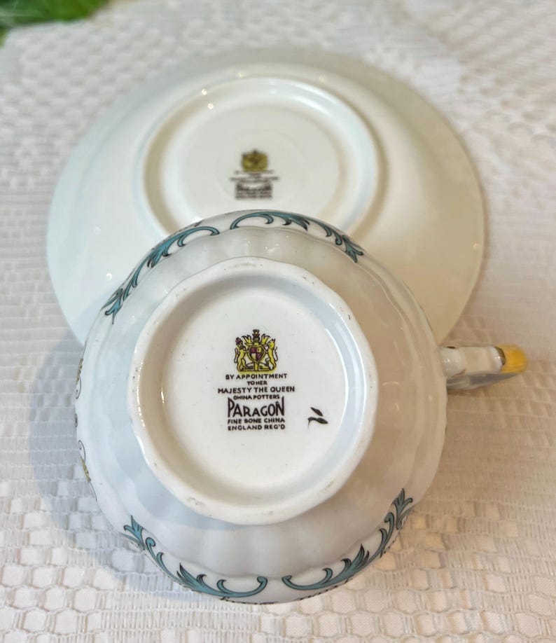 May include: White teacup and saucer with a blue and gold floral pattern. The bottom of the teacup has the text "By Appointment to Her Majesty The Queen China Potters Paragon Fine Bone China England Reg'd".
