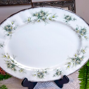 Pode incluir: Travessa oval branca para servir com borda prateada e design floral de rosas brancas e folhas verdes.