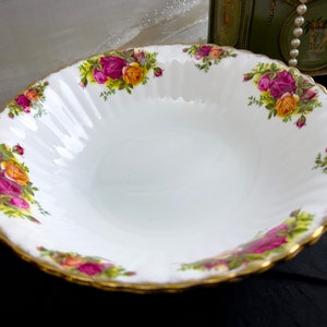 May include: A white ceramic bowl with a gold rim and a floral pattern featuring pink, yellow, and green roses. The bowl is designed with a scalloped edge.