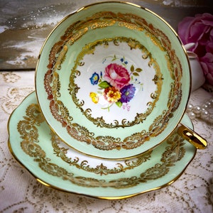 May include: A vintage teacup and saucer set. The cup and saucer are a soft green with gold trim and ornate gold filigree designs. The inside of the cup features a floral bouquet with pink, blue, and yellow flowers.