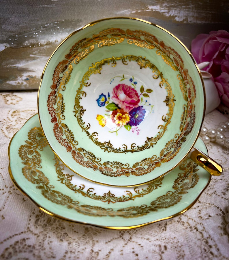 May include: A vintage teacup and saucer set. The cup and saucer are a soft green with gold trim and ornate gold filigree designs. The inside of the cup features a floral bouquet with pink, blue, and yellow flowers.
