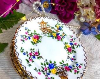 Könnte beinhalten: Ein Stapel weißer Porzellanteller mit goldenen Rändern und einem floralen Design. Die Teller weisen ein farbenfrohes Blumenmuster mit rosa, gelben und blauen Blumen sowie eine goldene Pagodenillustration auf. Die Teller sind auf einer weißen Spitzentischdecke gestapelt.