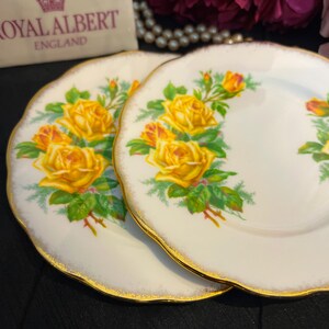 Puede incluir: Dos platos blancos Royal Albert con un diseño floral de rosas amarillas y borde dorado. Los platos tienen un borde festoneado y están sobre un fondo oscuro. El logotipo de Royal Albert es visible en el fondo.