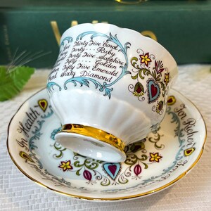 May include: A white teacup and saucer with a gold trim. The teacup has a floral design and the words "Thirty Five Coral, Forty Ruby, Forty Five Sapphire, Fifty Golden, Fifty Five Emerald, Sixty Diamond" written on it. The saucer has the words "In Health" and "For Richer" written on it.