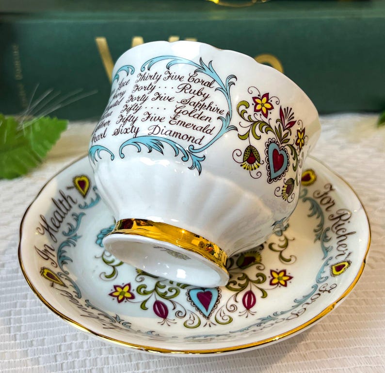 May include: A white teacup and saucer with a gold trim. The teacup has a floral design and the words "Thirty Five Coral, Forty Ruby, Forty Five Sapphire, Fifty Golden, Fifty Five Emerald, Sixty Diamond" written on it. The saucer has the words "In Health" and "For Richer" written on it.