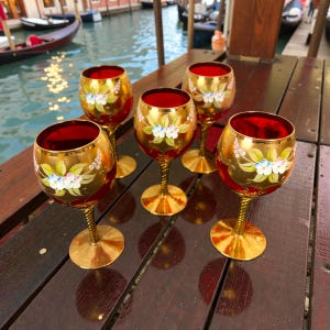 以下が含まれることがあります： 金色の縁と脚、赤い内側、白、青、緑の手描きの花柄が特徴の、5つの装飾的なワイングラス。 グラスは、光を反射する濃い木製の表面に配置されています。