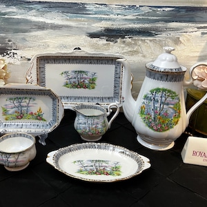 May include: A Royal Albert tea set in the Birchwood pattern. The set includes a teapot, creamer, sugar bowl, and two serving platters. The pieces are white with a grey and gold trim and feature a floral design with birch trees.
