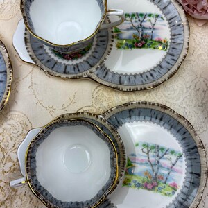 May include: A set of vintage teacups and saucers. The white cups and saucers have a gray and brown patterned border with gold trim. The saucers feature a landscape scene with trees and flowers.