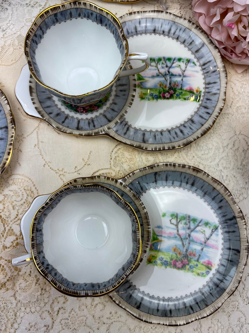 May include: A set of vintage teacups and saucers. The white cups and saucers have a gray and brown patterned border with gold trim. The saucers feature a landscape scene with trees and flowers.