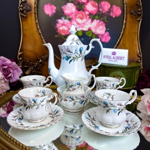 May include: A Royal Albert tea set featuring a white teapot, cups, saucers, and a creamer. The set is decorated with a floral pattern in blue, purple, and green. The set is displayed on a mirrored surface with a gold-colored frame.