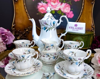 Könnte beinhalten: Ein Royal Albert Teeservice mit weißer Teekanne, Tassen, Untertassen und Sahnekännchen. Das Set ist mit einem floralen Muster in Blau, Lila und Grün verziert. Präsentiert auf einer verspiegelten Oberfläche mit goldenem Rahmen.