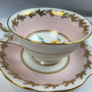 Puede incluir: Un juego de taza y platillo de t&eacute; rosa con ribete dorado y dise&ntilde;o floral. La taza y el platillo est&aacute;n decorados con un delicado patr&oacute;n floral en tonos amarillos, azules y verdes.