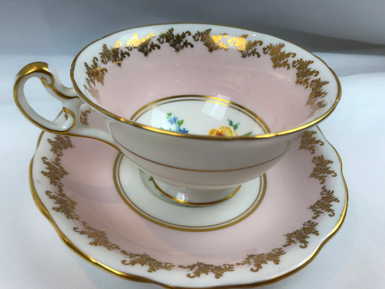 Puede incluir: Un juego de taza y platillo de t&eacute; rosa con ribete dorado y dise&ntilde;o floral. La taza y el platillo est&aacute;n decorados con un delicado patr&oacute;n floral en tonos amarillos, azules y verdes.