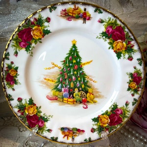May include: A white porcelain plate with a gold rim and a Christmas tree design. The plate features a decorated green Christmas tree, wrapped gifts, and a border of red and yellow roses with green leaves. A festive holiday plate.