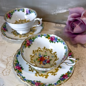 Peut inclure: Deux tasses et soucoupes blanches avec des motifs floraux. Les tasses et les soucoupes ont des bordures dorées et un motif de fleurs roses, bleues et jaunes. Une rose pourpre est en arrière-plan.