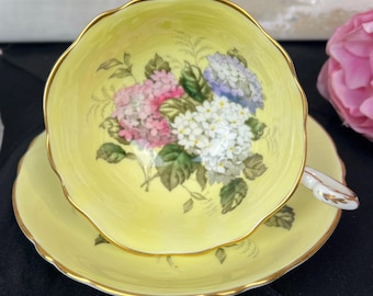 May include: A yellow teacup and saucer with a floral design featuring pink, white, and purple hydrangeas. The cup and saucer have a gold trim.