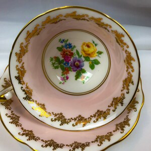 Puede incluir: Una taza de t&eacute; y un platillo rosas con un borde dorado y un dise&ntilde;o floral. Las flores son amarillas, moradas, rosas y azules.