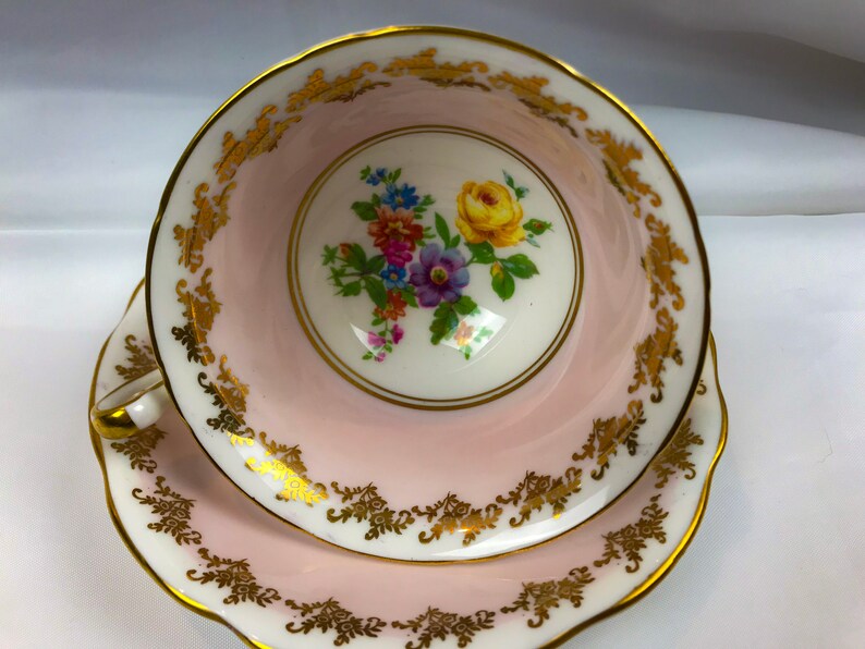 Puede incluir: Una taza de t&eacute; y un platillo rosas con un borde dorado y un dise&ntilde;o floral. Las flores son amarillas, moradas, rosas y azules.