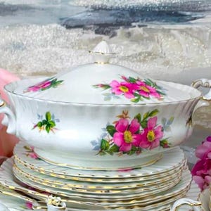 Op de afbeelding: Een witte porseleinen theeset met een bloemenpatroon met roze rozen. De set bevat een gedekte serveerschaal, borden en een theekopje. De set is versierd met een gouden rand.