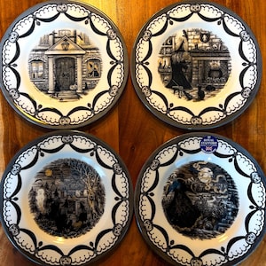 May include: Set of four black and white Halloween themed dinner plates with a silver rim. The plates feature illustrations of a haunted house, a witch, a graveyard, and a spooky tree.
