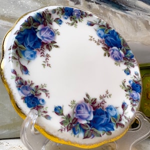 Może przedstawiać: Biały talerz z falistym brzegiem i złotą obwódką, ozdobiony wzorem niebieskich róż i zielonych liści. Kwiatowy wzór rozmieszczony jest wokół krawędzi talerza. Talerz jest prezentowany na przezroczystym stojaku.