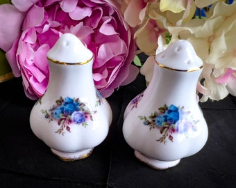 May include: A pair of white ceramic salt and pepper shakers with a floral design featuring blue roses. The shakers have a gold trim and a classic, elegant shape, perfect for a dining table setting.